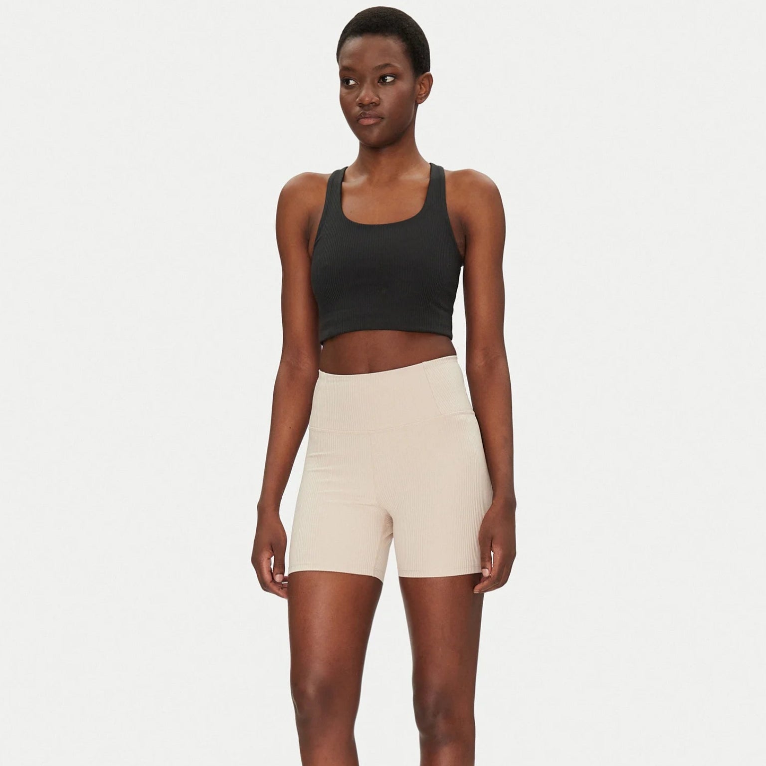 Eine Frau steht vor einem schlichten weißen Hintergrund und trägt einen schwarzen Sport-BH, hellbeige Biker-Shorts und weiße Turnschuhe. Sie hat kurzes Haar und eine entspannte, selbstbewusste Körperhaltung, mit den Armen an den Seiten.