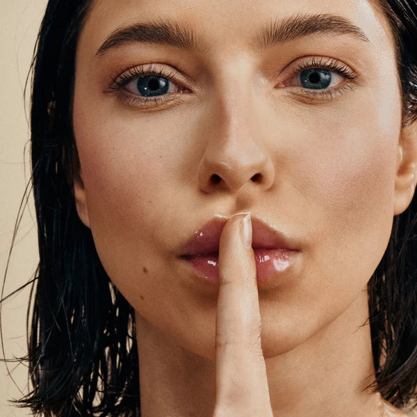 Eine Frau mit nassem, dunklem Haar hält ihren Zeigefinger an die Lippen, um Ruhe zu signalisieren. Sie hat klare Haut, ist minimal geschminkt und blickt mit einem ruhigen, ernsten Blick direkt in die Kamera.