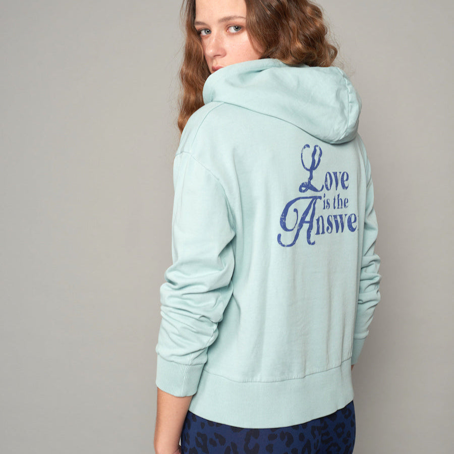 Frau trägt einen hellblauen Hoodie mit der Aufschrift "Love is the Answer" auf dem Rücken, sieht über die Schulter.