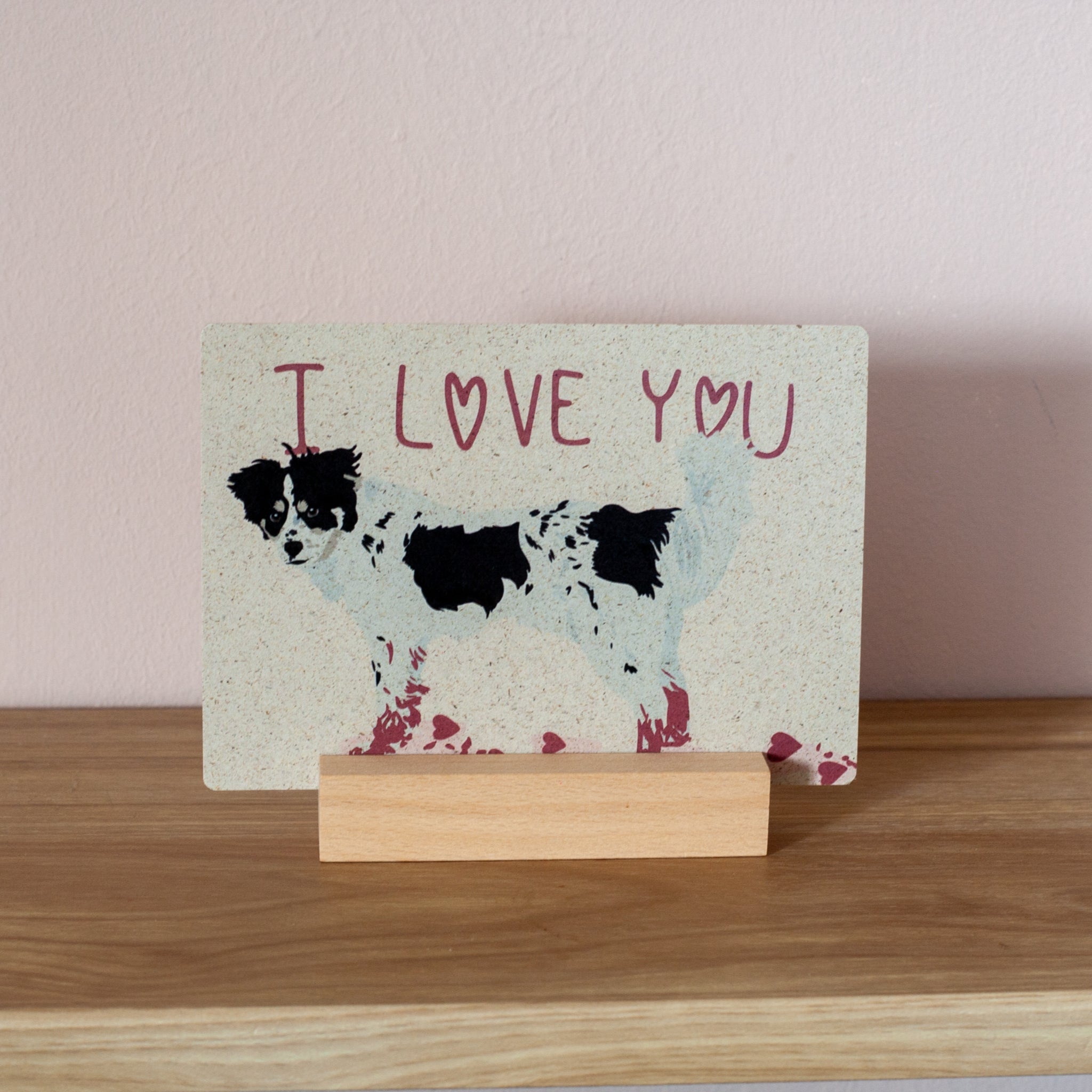 Eine Karte mit der Illustration eines schwarz-weißen Hundes und den Worten ICH LIEBE DICH in rosa Schrift darüber, auf einem Holzständer vor einer hellrosa Wand.