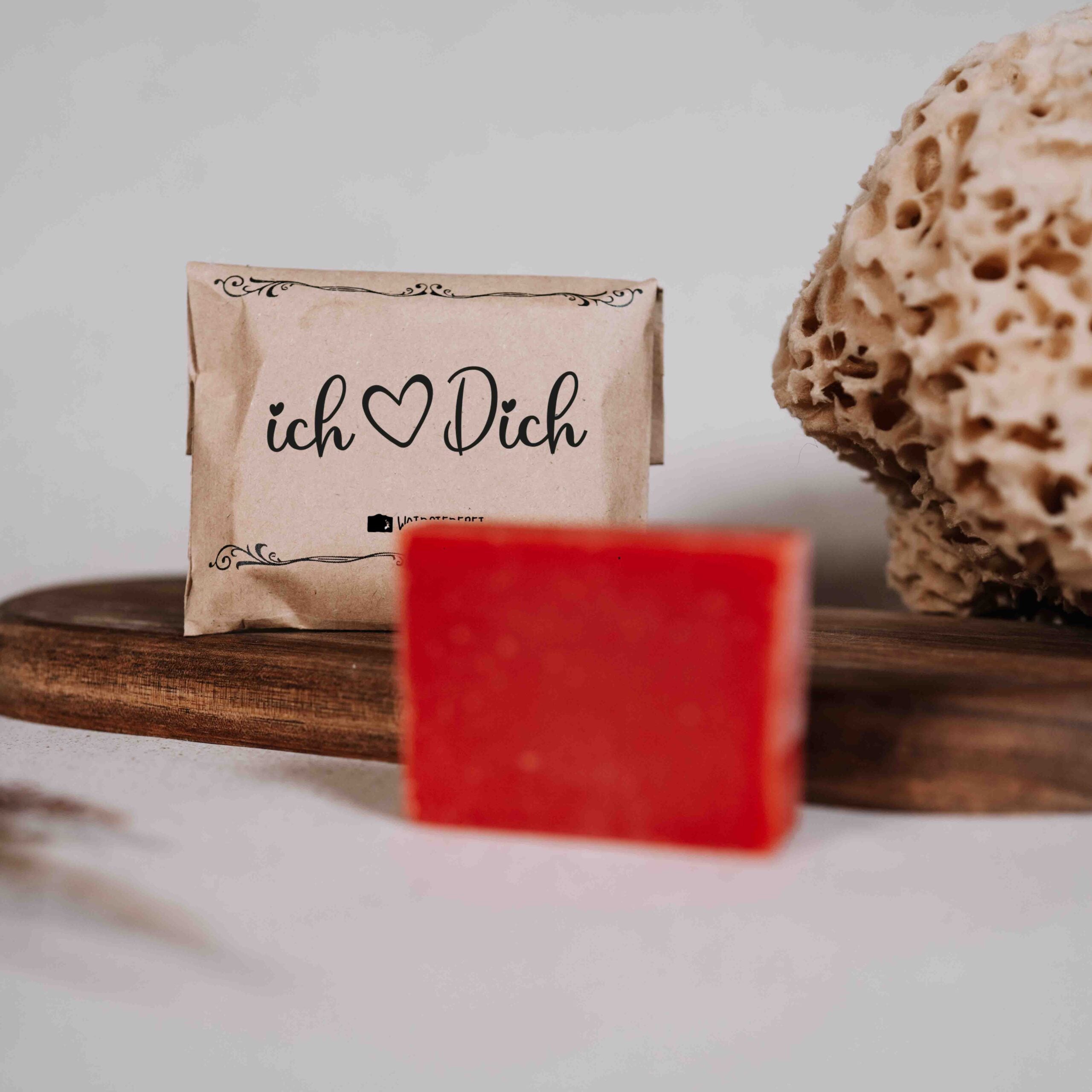 Ein Stück rote Seife liegt auf einem Holztablett neben einem Naturschwamm. Dahinter ist ein Papierpaket mit der Aufschrift "Ich ♥ Dich" vor einem hellen Hintergrund zu sehen.