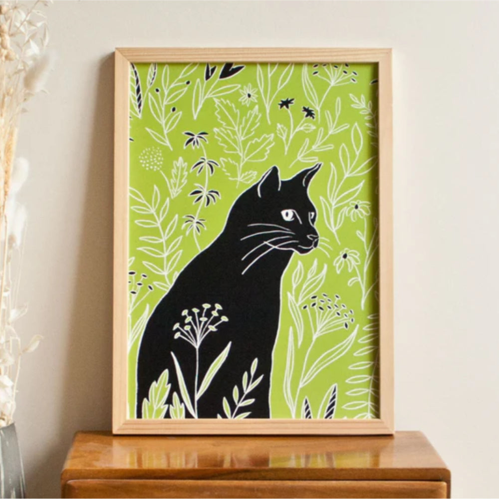 Eine gerahmte Illustration einer schwarzen Katze, umgeben von grünen Blattpflanzen und weißen Blumenakzenten vor einem hellgrünen Hintergrund, befindet sich auf einer Holzoberfläche.