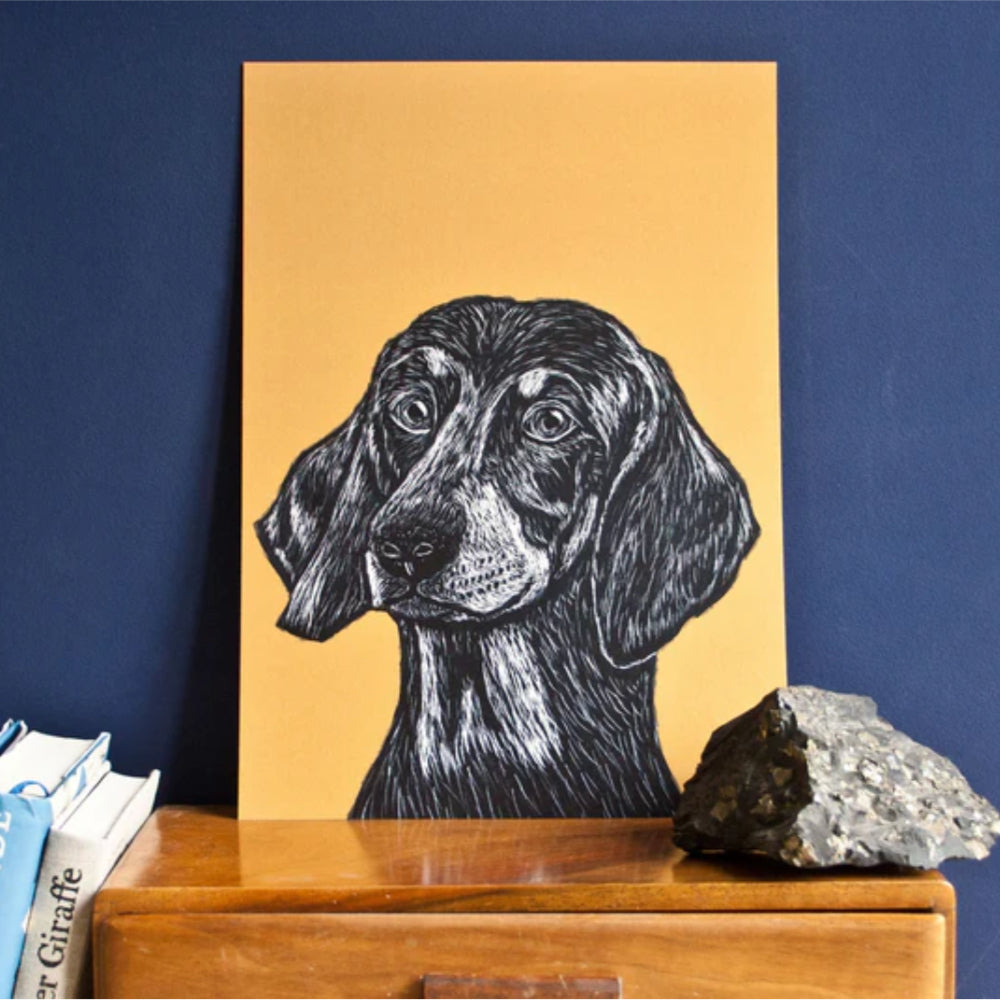 Eine Schwarz-Weiß-Zeichnung eines Hundes mit Schlappohren auf hellbraunem Papier ist an eine dunkelblaue Wand gelehnt und liegt auf einer Holzkommode neben einem Stein und einem Stapel Bücher.