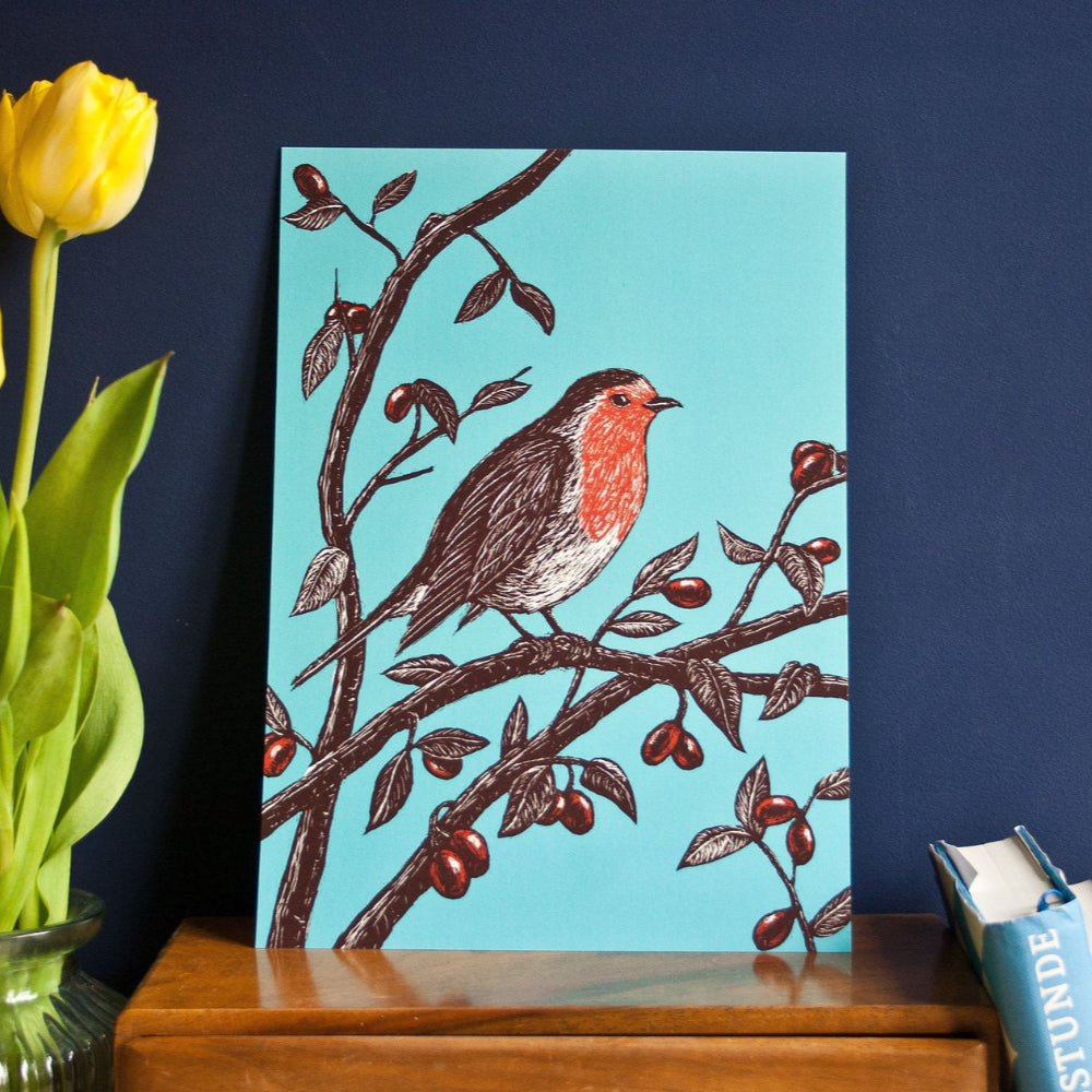 Eine farbenfrohe Illustration eines Rotkehlchens, das auf einem Zweig mit roten Beeren sitzt, vor einem hellblauen Hintergrund. Eine gelbe Tulpe in einer Vase und gestapelte Bücher stehen in der Nähe auf einer Holzfläche.