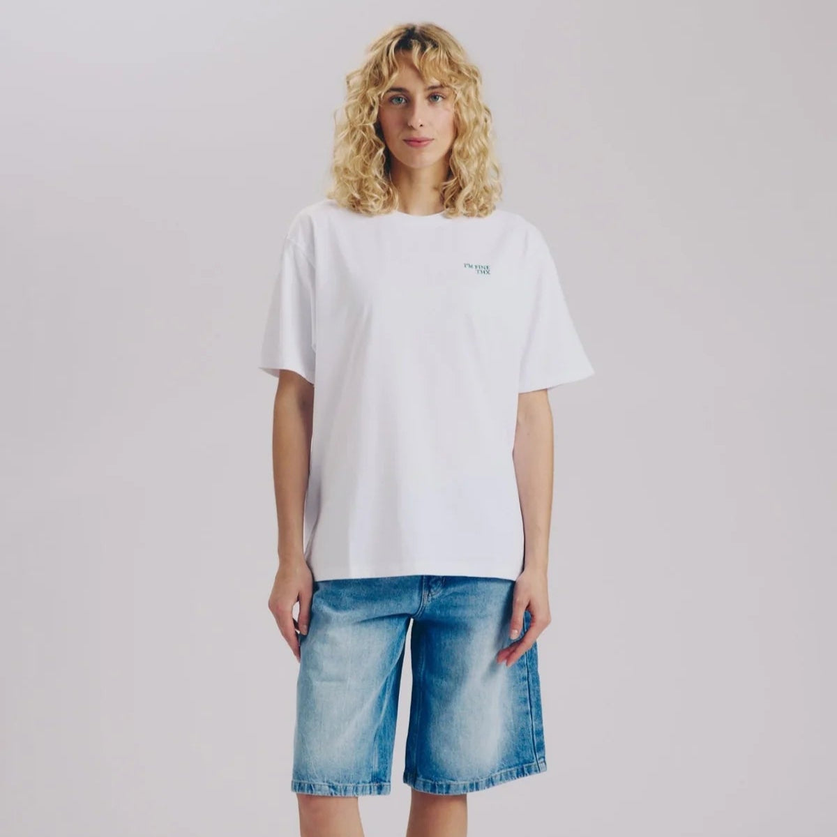 Eine Person mit lockigem blondem Haar, die ein einfaches weißes T-Shirt, knielange blaue Jeans-Shorts, weiße Socken und weiße Turnschuhe trägt, steht vor einem einfachen hellen Hintergrund.