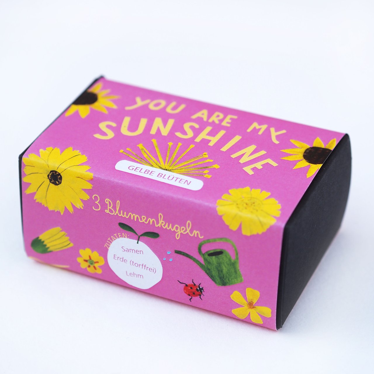 Eine kleine Schachtel mit einem leuchtend rosa Design, verziert mit gelben Blumen, einer Gießkanne, einem Marienkäfer und dem Text Du bist mein Sonnenschein und 3 Blumenkugeln auf Deutsch. Die Schachtel enthält gelbe Blumensamen.