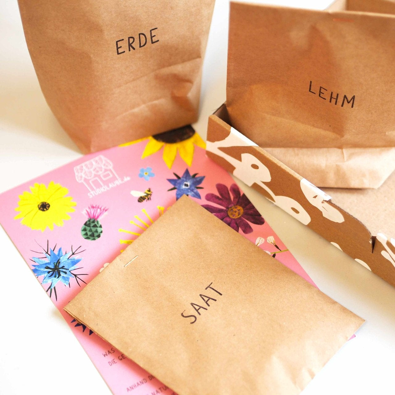 Drei braune Papiertüten mit den Aufschriften ERDE (Erde), LEHM (Lehm) und SAAT (Samen) sind mit einer blumengemusterten Schachtel und einem rosafarbenen Blatt mit farbenfrohen Blumenabbildungen angeordnet.