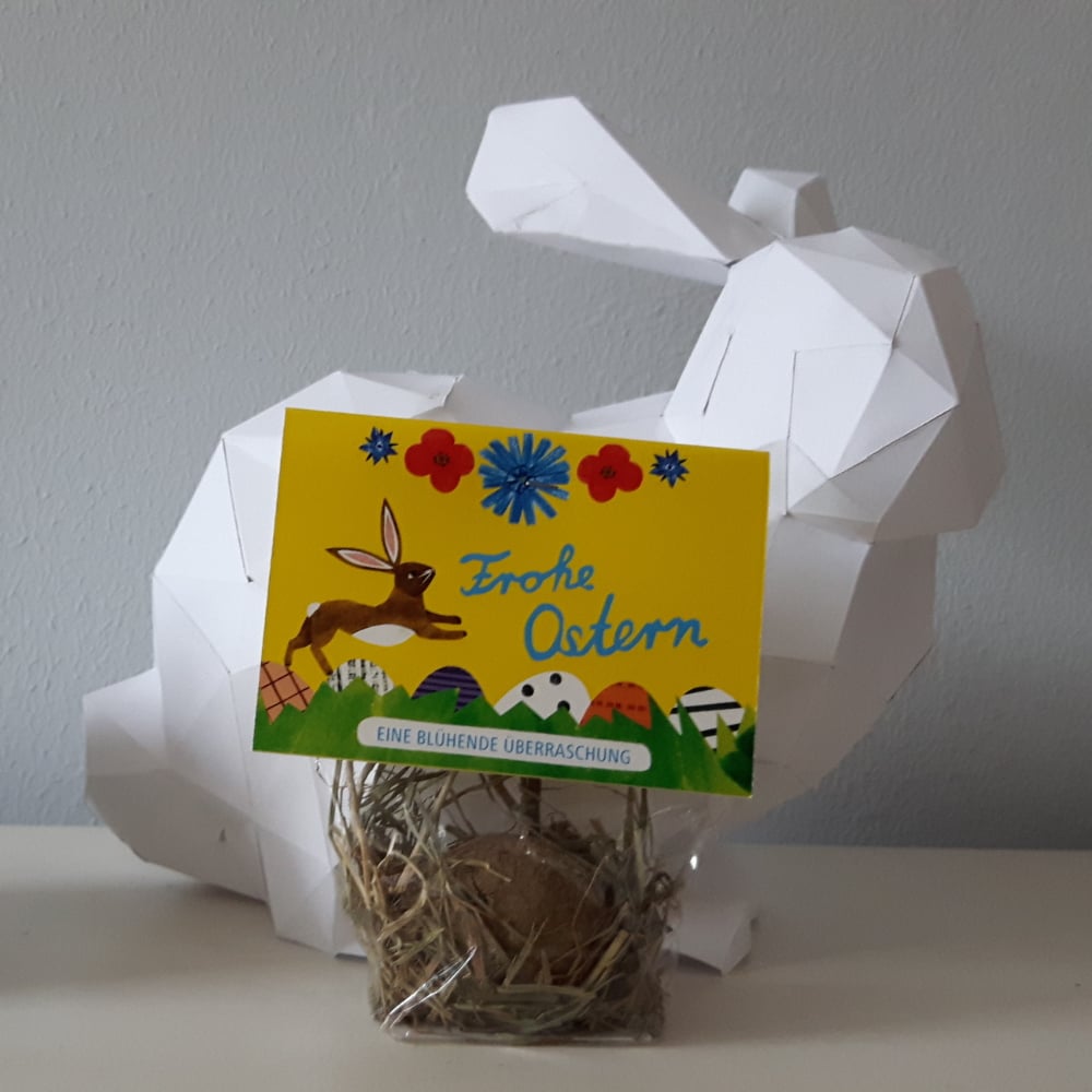 Ein geometrischer weißer Papierhase sitzt hinter einer kleinen Tüte mit Heu und einem braunen Ei darin. An der Tüte ist eine bunte Karte mit der Aufschrift "Frohe Ostern" und österlichen Dekorationen befestigt.