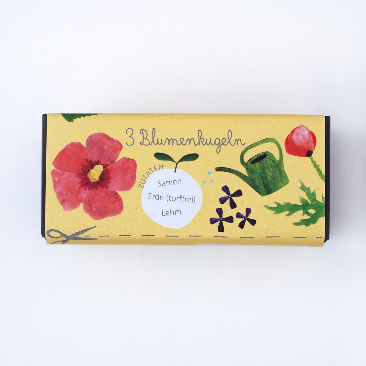 Eine gelbe Schachtel mit bunten Zeichnungen einer roten Blume, lila Blumen, einer grünen Gießkanne und Blättern. Auf der Schachtel steht "3 Blumenkugeln" und die Zutaten sind aufgelistet: "Samen, Erde (torffrei), Lehm".