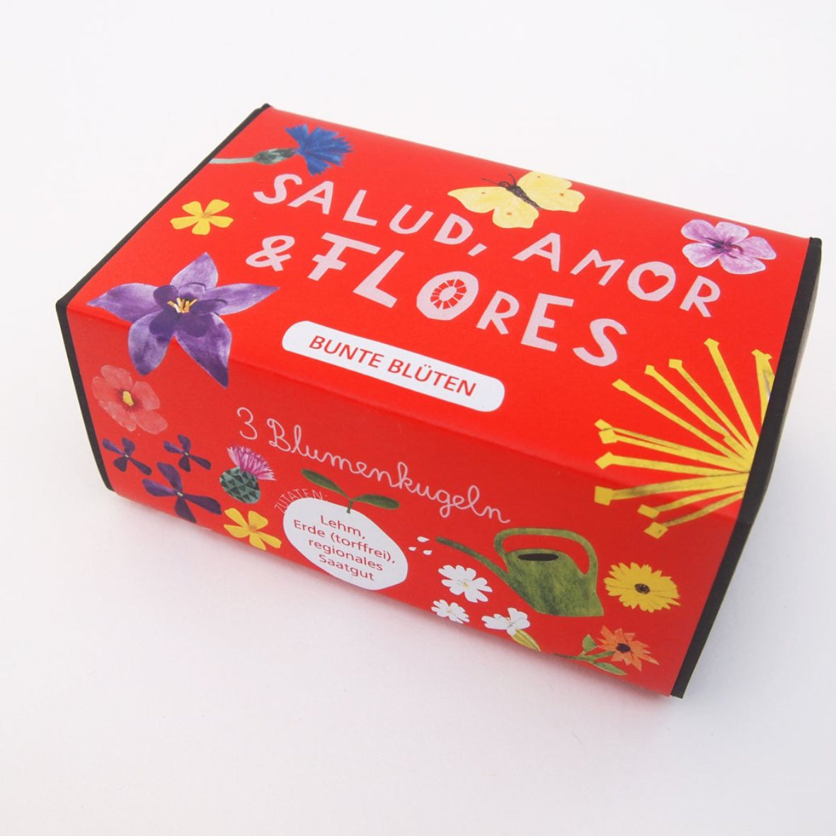 Eine rechteckige rote Schachtel mit bunten Blumenillustrationen und dem Text "SALUD, AMOR & FLORES" zusammen mit "BUNTE BLÜTEN" und "3 Blumenkugeln". Eine kleine grüne Gießkanne und ein Blumensymbol sind ebenfalls zu sehen.
