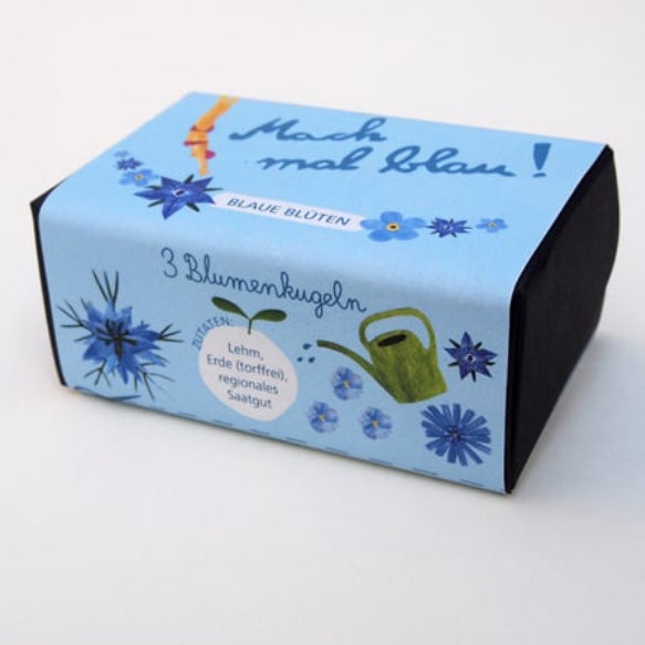 Eine rechteckige Schachtel mit einem blauen Etikett mit blauen Blumen, einer Gießkanne und einem deutschen Text, der "Mach mal Blau!" und "3 Blumenkugeln" lautet. Die Schachtel scheint Blumenkugeln zu enthalten.