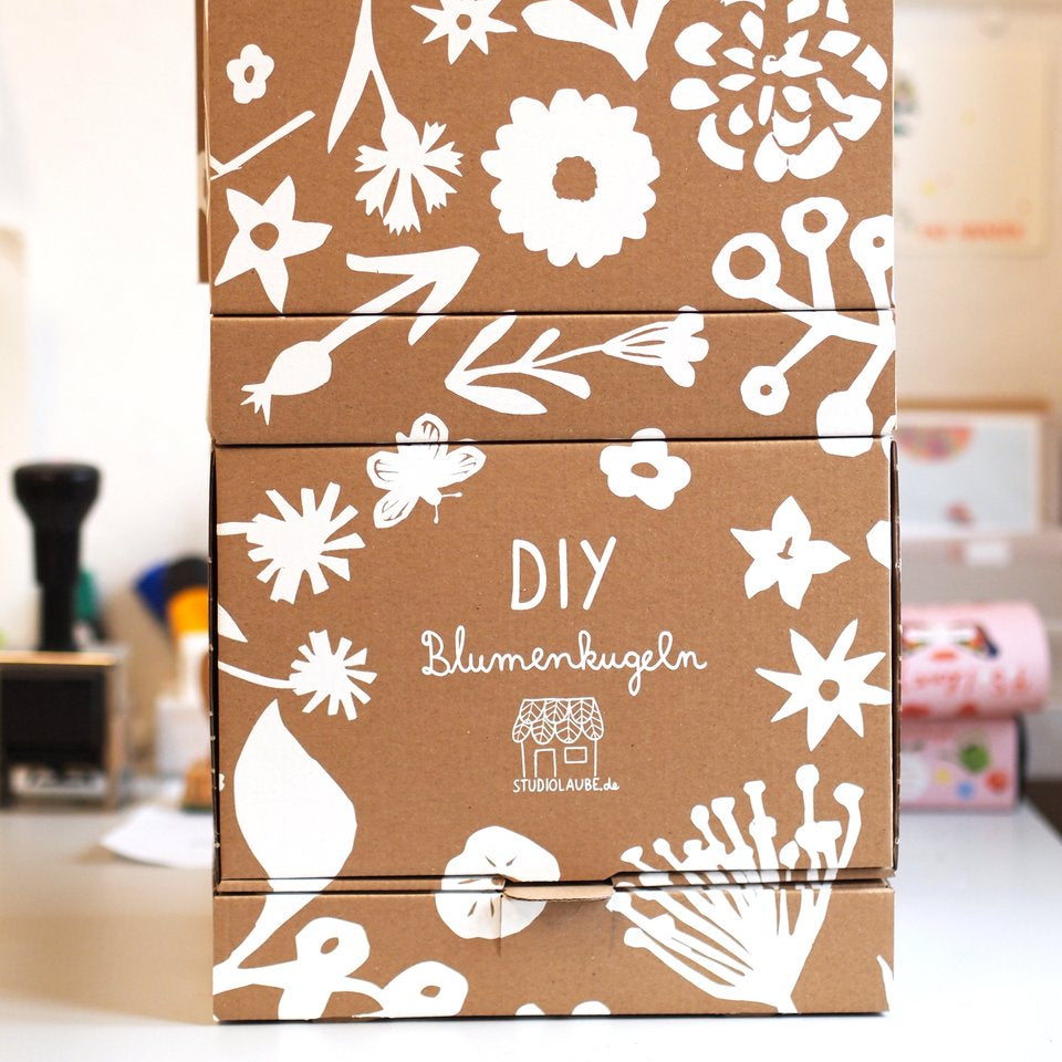 Drei gestapelte braune Kartons mit weißen Blumen- und Blattillustrationen. Auf der mittleren Schachtel steht DIY Blumen kugeln mit einem kleinen Hauslogo darunter. Der Hintergrund ist eine sanft fokussierte Innenumgebung.
