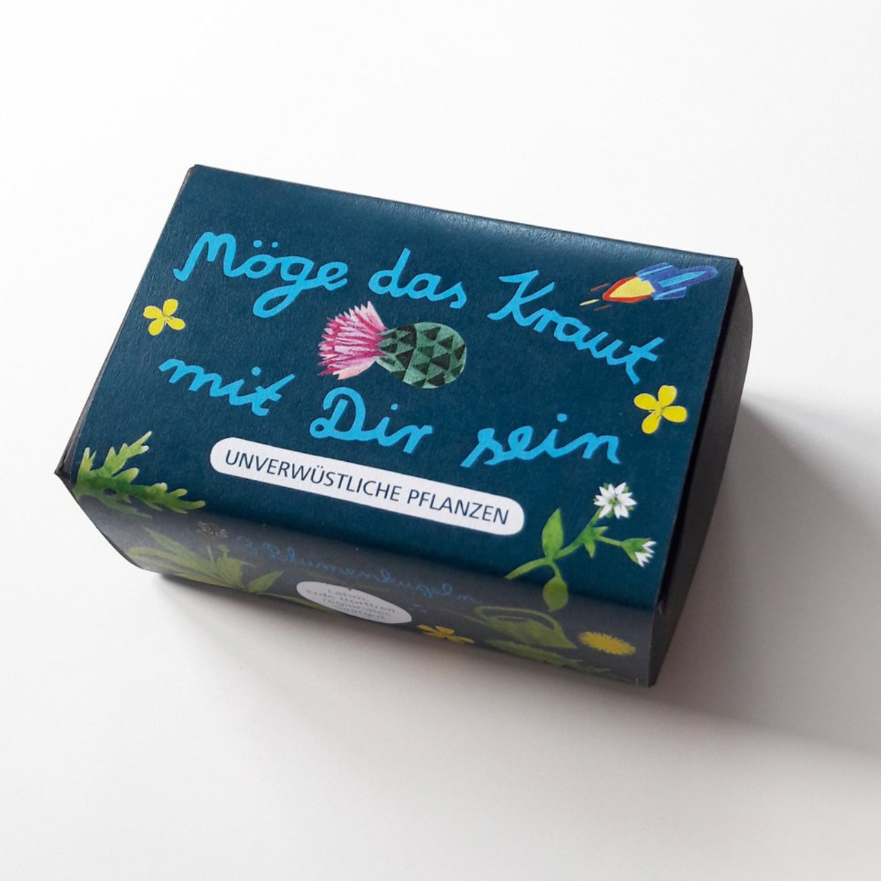 Eine dunkelblaue Schachtel mit floralen Illustrationen, deutschem Text Möge das Kraut mit Dir sein und UNVERWÜSTLICHE PFLANZEN auf dem Deckel, dargestellt auf einer weißen Fläche.