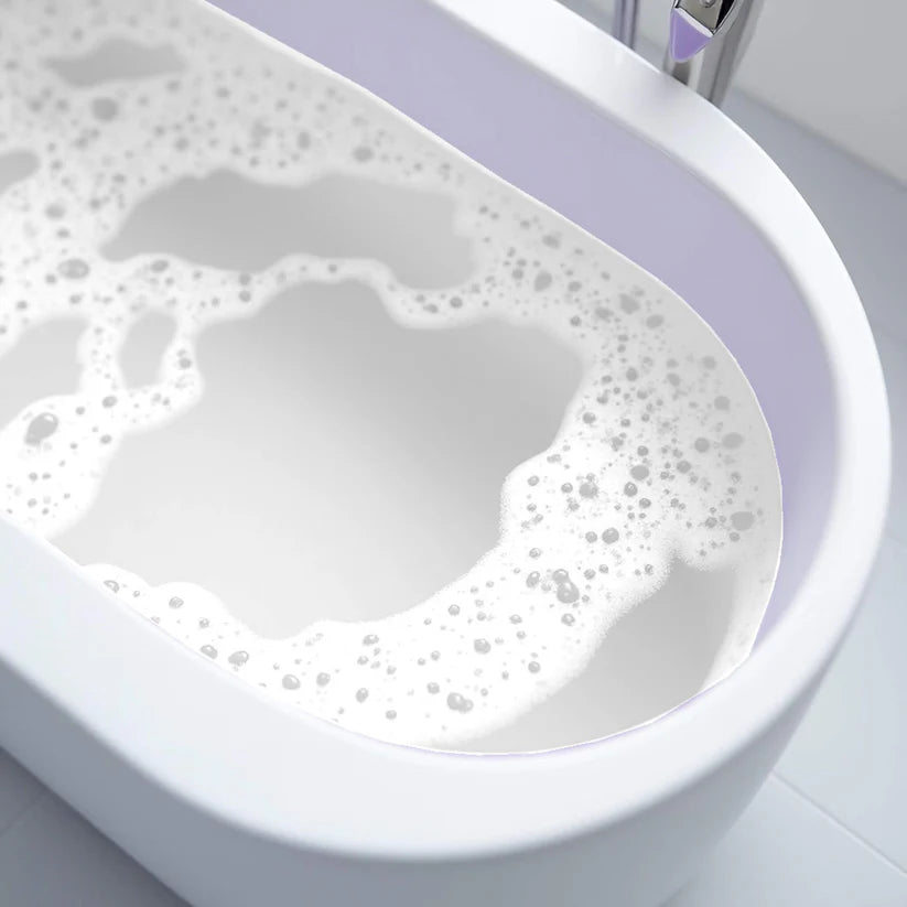Nahaufnahme einer weißen Badewanne, die mit Wasser und weißen, schaumigen Blasen gefüllt ist. Die Wanne ist oval und steht auf einem hellen Kachelboden.