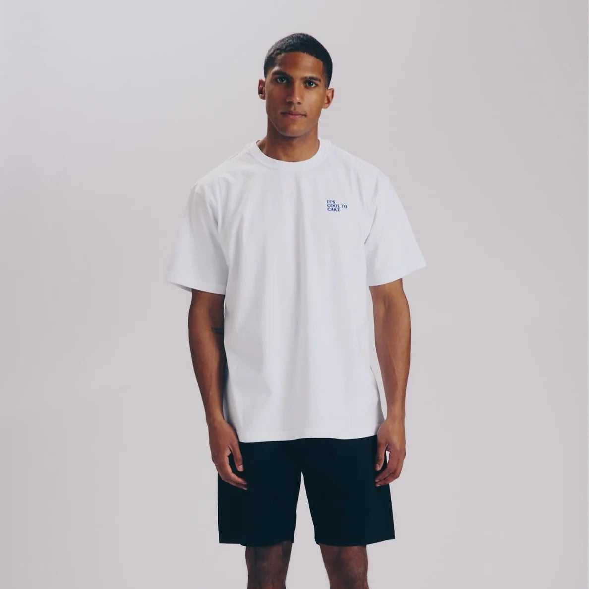 Ein Mann steht vor einem schlichten Hintergrund und trägt ein weißes T-Shirt, schwarze Shorts, weiße Socken und weiße Turnschuhe. Er blickt mit neutralem Gesichtsausdruck und entspannter Körperhaltung nach vorne.