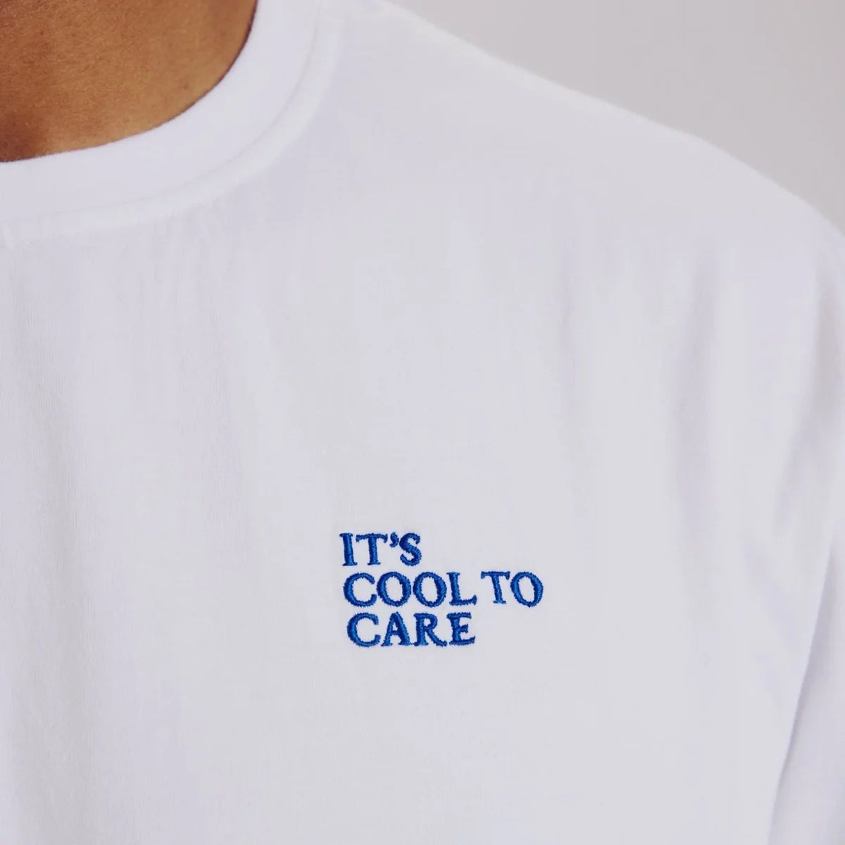Nahaufnahme eines weißen T-Shirts mit blauem gesticktem Text: "IT'S COOL TO CARE". Der obere Teil der Brust und des Halses einer Person ist zu sehen.