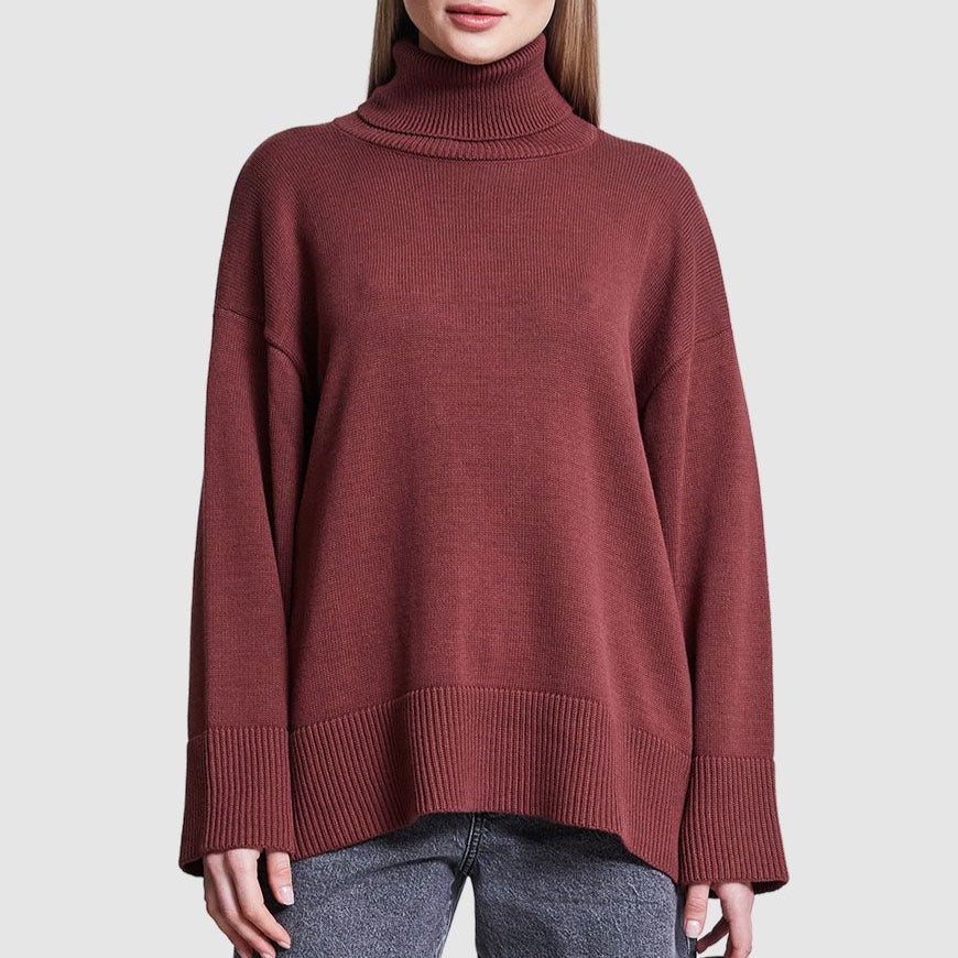 Eine Person, die einen weiten, langärmeligen, kastanienbraunen Rollkragenpullover mit gerippten Bündchen und Saum trägt, kombiniert mit einer grauen Jeans, steht vor einem einfarbigen hellgrauen Hintergrund.