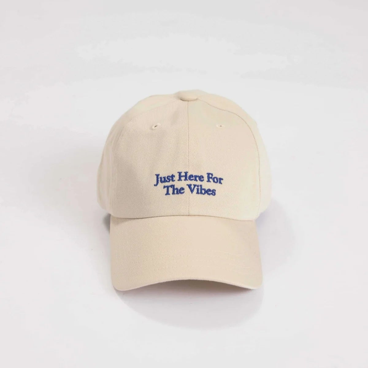 Eine beigefarbene Baseballkappe mit dem blau aufgestickten Schriftzug Just Here For The Vibes auf der Vorderseite vor einem schlichten weißen Hintergrund.