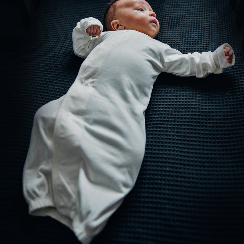 Ein Baby in einem weißen Schlafanzug liegt auf einer dunklen Decke.