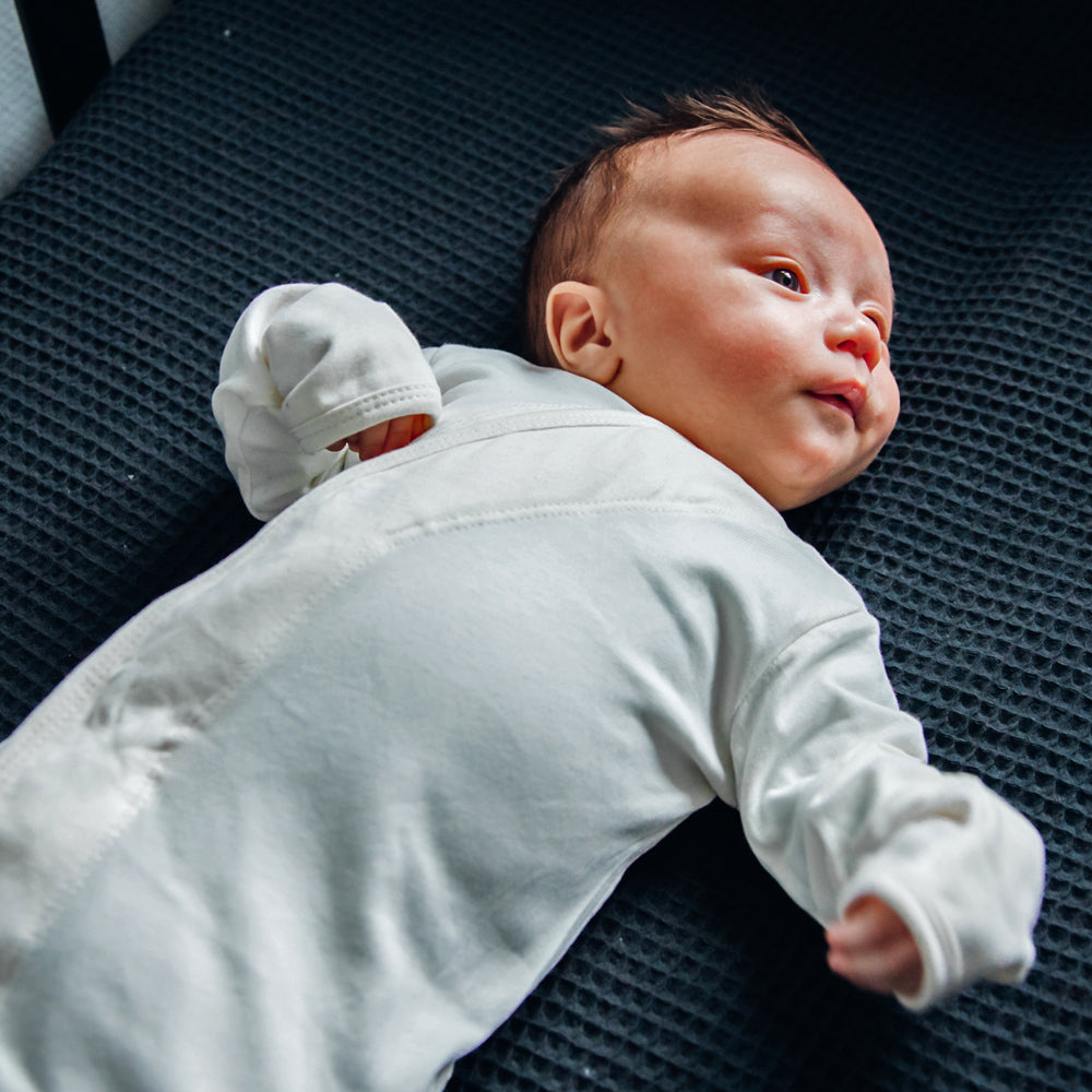 Ein Baby in einem weißen Strampler liegt auf einer dunkelgrauen Decke und blickt zur Seite.