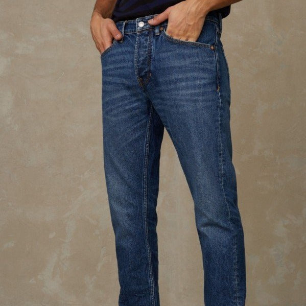 Person in blauer Jeans mit Händen in den Hosentaschen vor beigem Hintergrund.