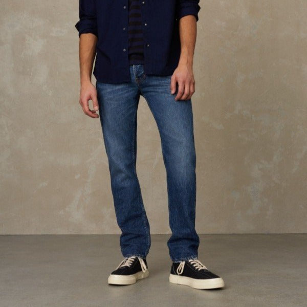 Person in blauer Jeans und Sneakers steht vor einer beigefarbenen Wand, Arme entspannt an den Seiten.