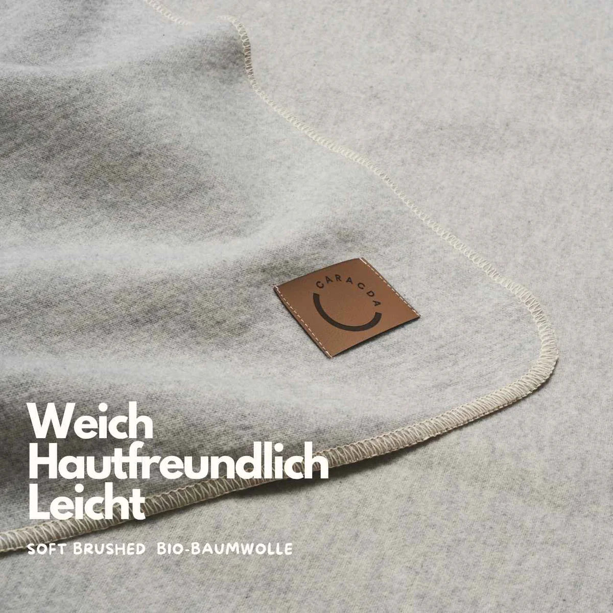 Nahaufnahme einer weichen, hellgrauen Stoffdecke mit beigen Nähten und einem braunen Etikett mit der Aufschrift GARAPAPA. Text auf dem Bild: Weich Hautfreundlich Leicht. Weich gebürstete Bio-Baumwolle.