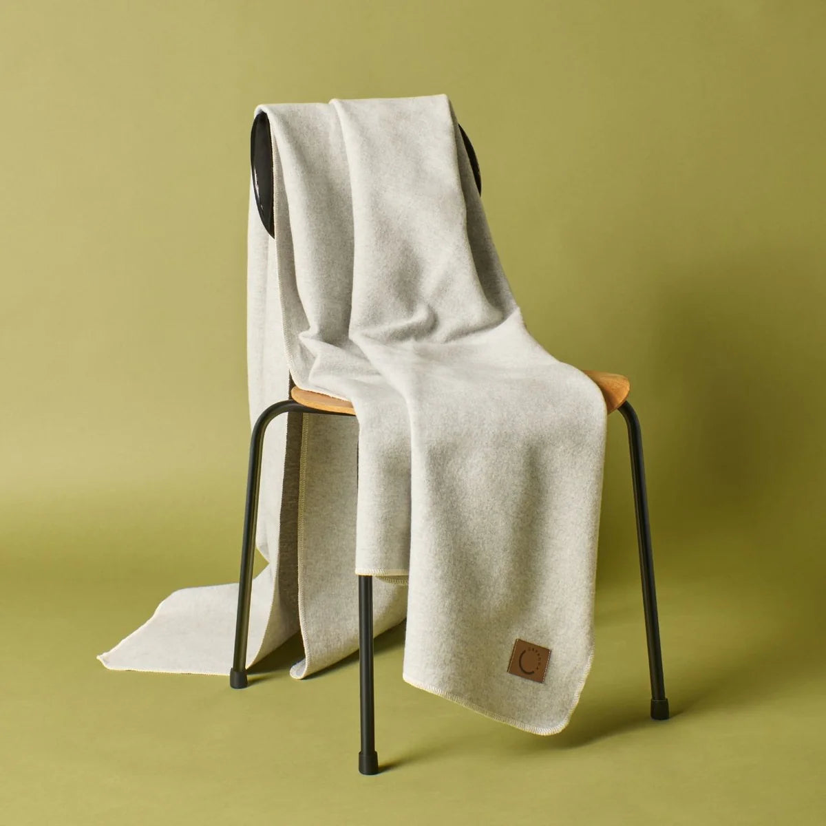 Eine weiche, hellgraue Decke mit einem braunen Lederaufnäher ist über die Rückenlehne und die Sitzfläche eines einfachen schwarzen Metallstuhls mit einer Holzsitzfläche vor einem olivgrünen Hintergrund drapiert.