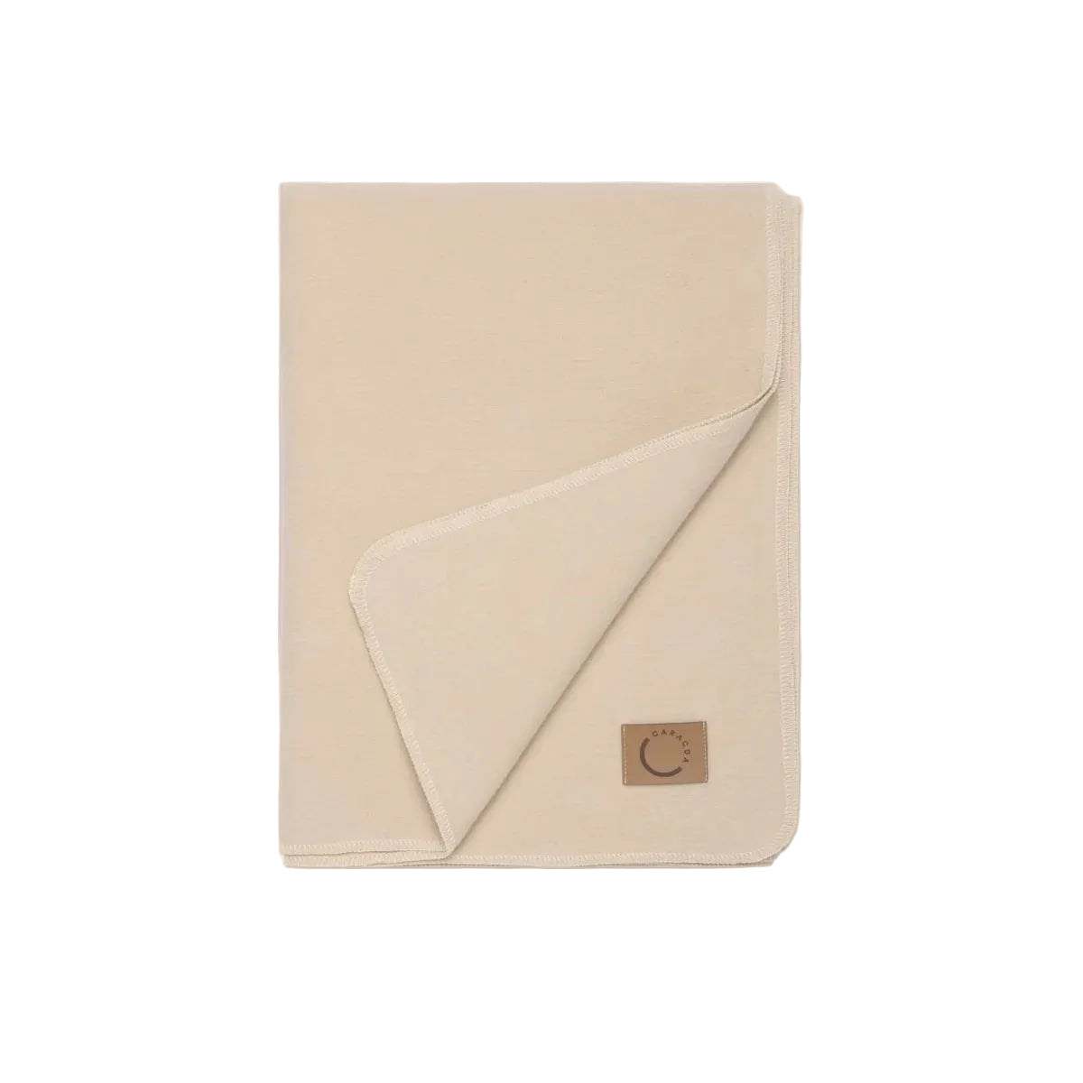 Eine ordentlich gefaltete beigefarbene Decke, bei der eine Ecke umgeschlagen ist und ein aufgenähtes braunes Etikett mit dem Motiv einer Mondsichel und Sonne zum Vorschein kommt. Die Decke ist vor einem passenden beigen Hintergrund ausgestellt.