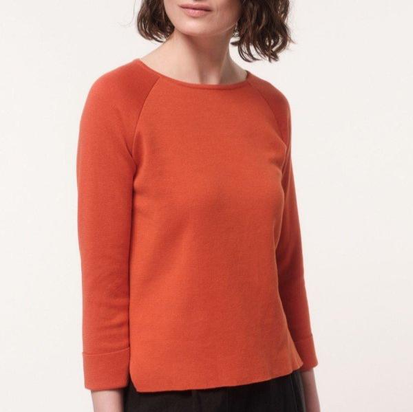Frau trägt einen orangefarbenen Pullover mit 3/4-Ärmeln vor einem weißen Hintergrund.