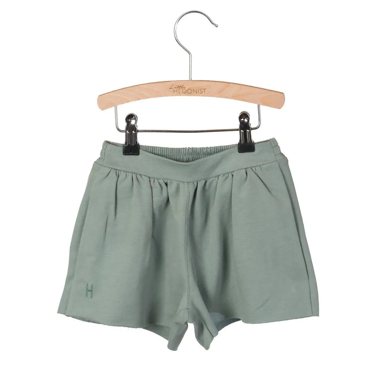 Hellgrüne Kindershorts mit elastischem Bund, ausgestellt auf einem Holzbügel mit der Aufschrift Little HEDONIST und befestigt mit zwei schwarzen Klammern.