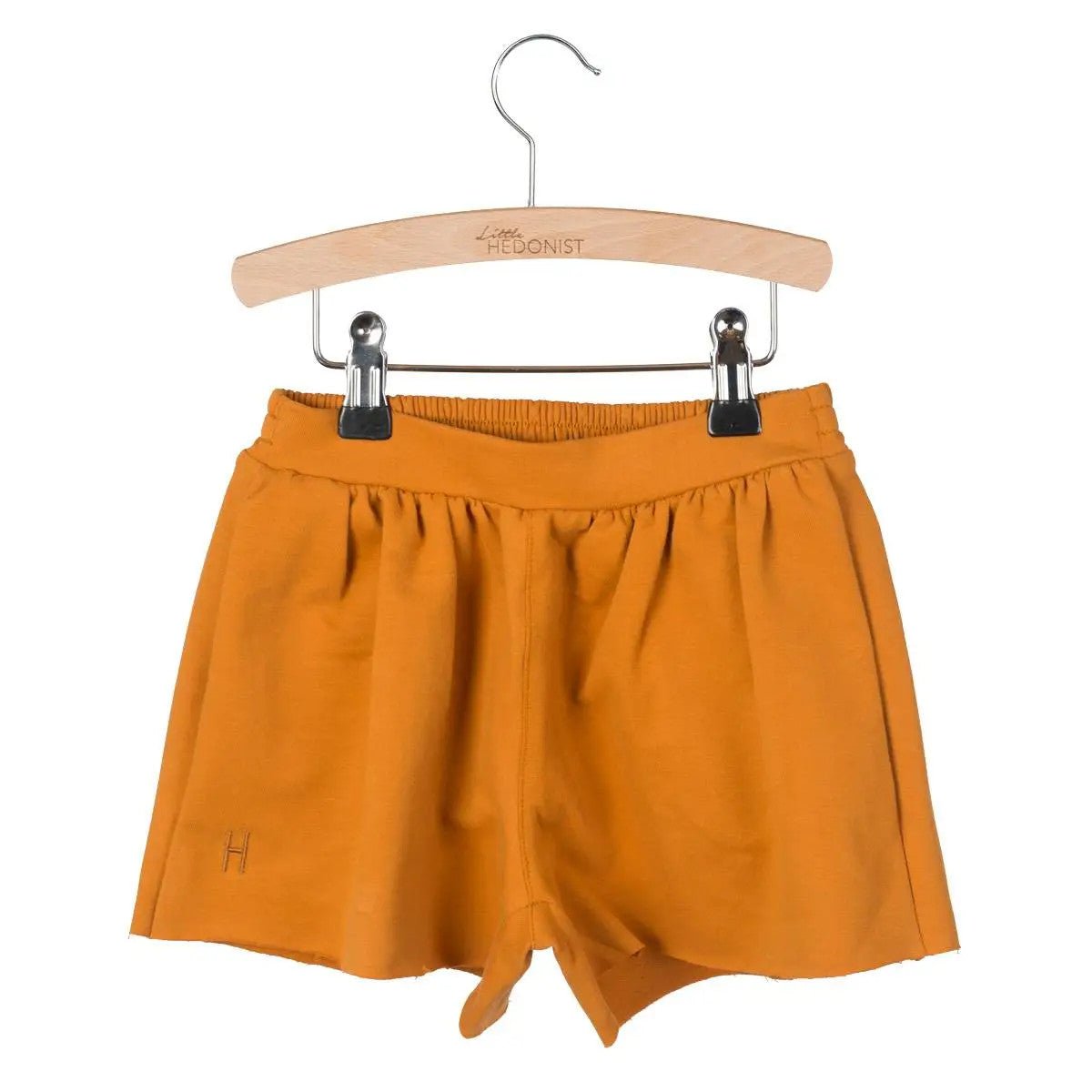 An einem Holzbügel mit der Aufschrift HEDONIST hängt ein Paar orangefarbene Shorts mit einem elastischen Bund. Die Shorts haben ein einfaches Design mit einem kleinen H, das in der Nähe des Saums aufgenäht ist.