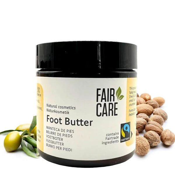 Ein Tiegel Fair Care Foot Butter mit fair gehandelten Zutaten steht neben Shea-Nüssen und Olivenfrüchten auf einem weißen Hintergrund. Das Etikett enthält Text in mehreren Sprachen und die Fairtrade-Zertifizierung.