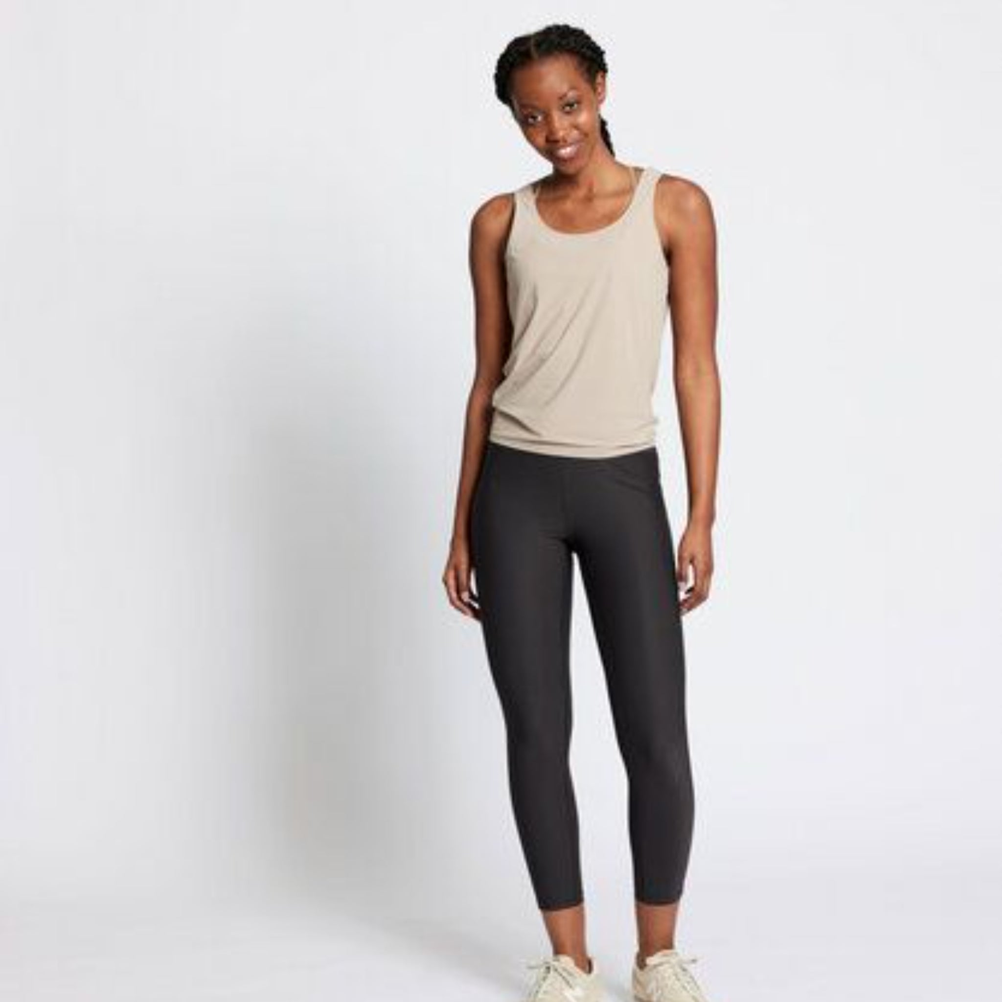 Eine Frau steht vor einem schlichten weißen Hintergrund und trägt ein beigefarbenes ärmelloses Oberteil, schwarze Leggings und helle Turnschuhe. Sie lächelt und hat die Arme locker an der Seite.