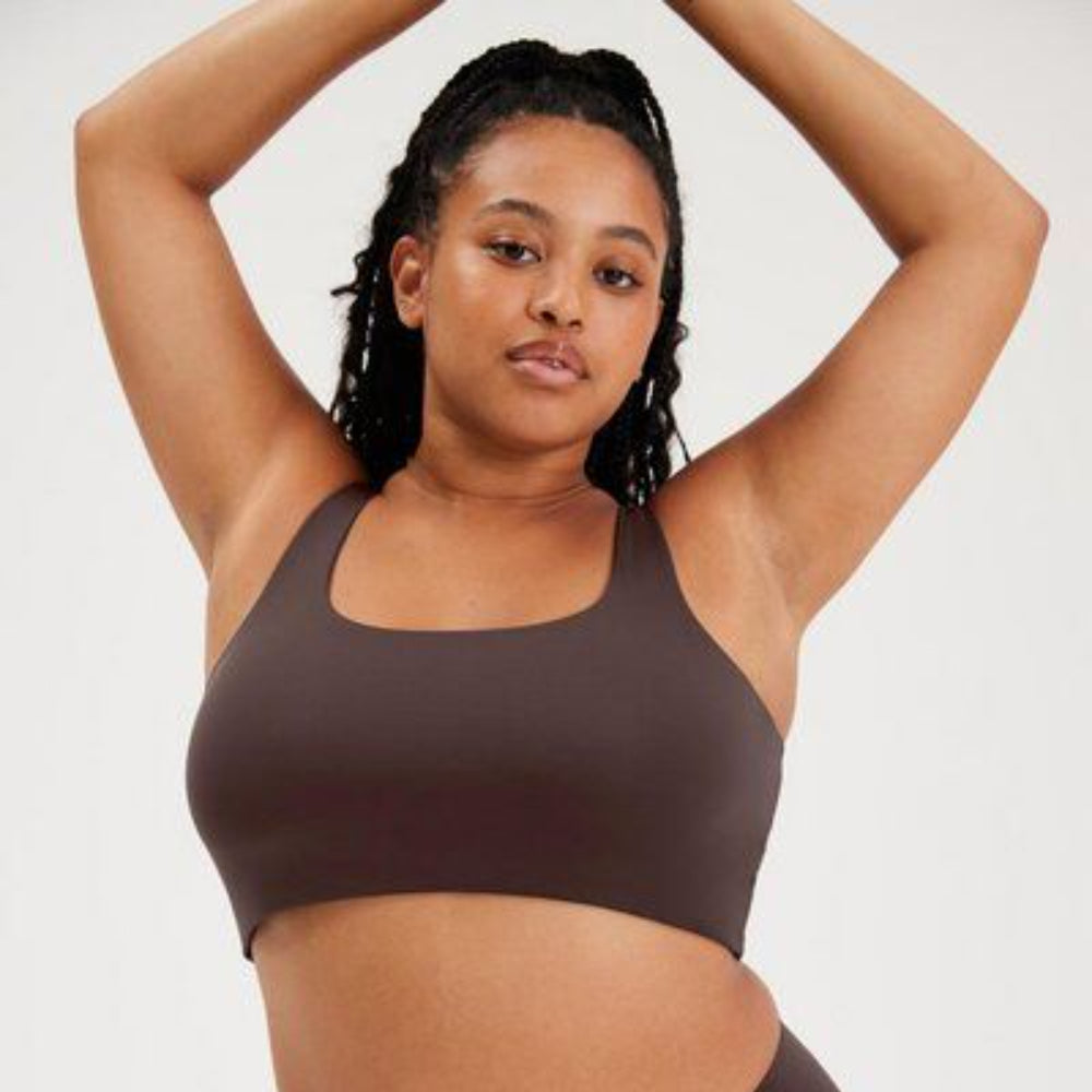 Eine Frau mit langen, lockigen Haaren in einem Pferdeschwanz trägt einen dunkelbraunen Sport-BH, hebt die Arme über den Kopf und blickt selbstbewusst in die Kamera vor einem hellen Hintergrund.