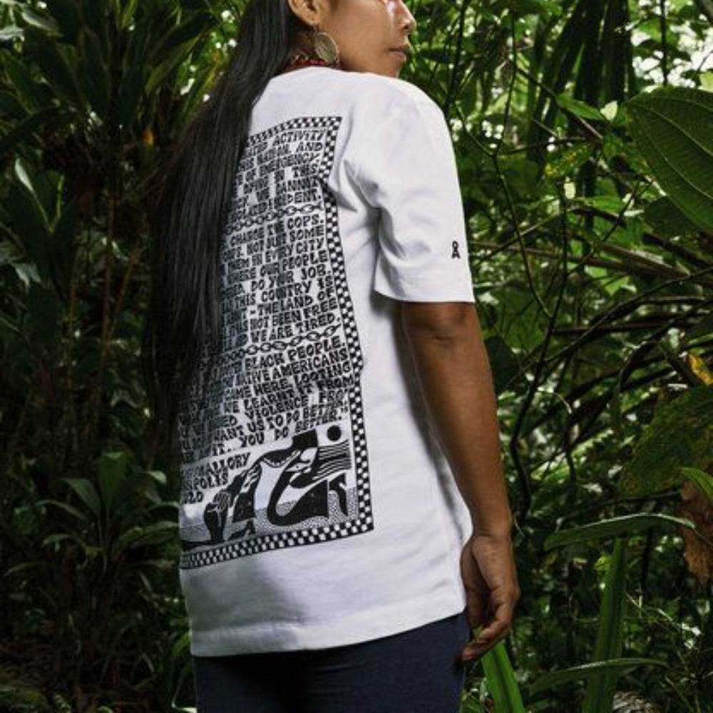 Eine Person steht in einem üppigen, grünen Wald und trägt ein weißes T-Shirt mit schwarzem grafischem Text und Mustern auf dem Rücken. Das Gesicht der Person ist abgewandt, und ihr langes Haar fällt in Kaskaden über die Schulter.