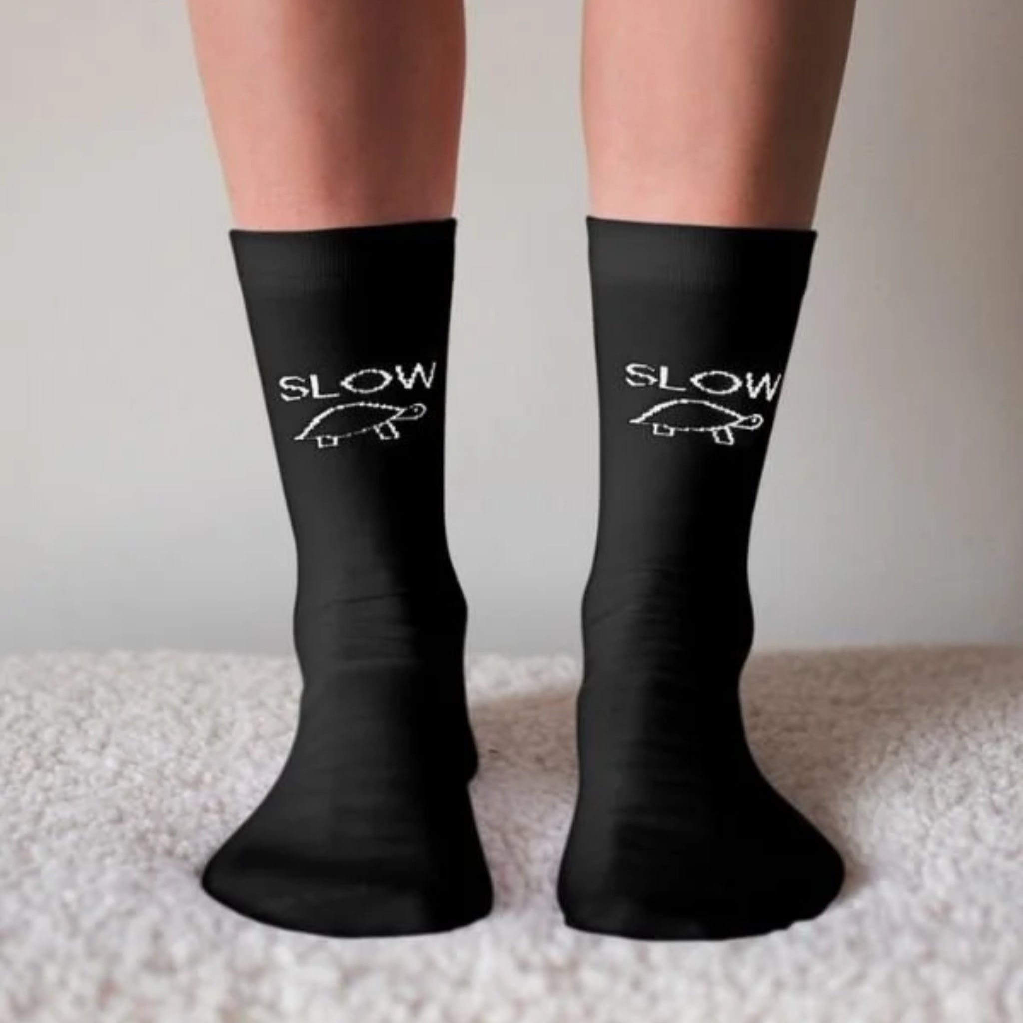 Ein Paar Füße in schwarzen Socken mit dem weißen Umriss einer Schildkröte und dem Wort "SLOW" darüber auf jeder Socke, die auf einem weichen, hellen Teppich stehen.