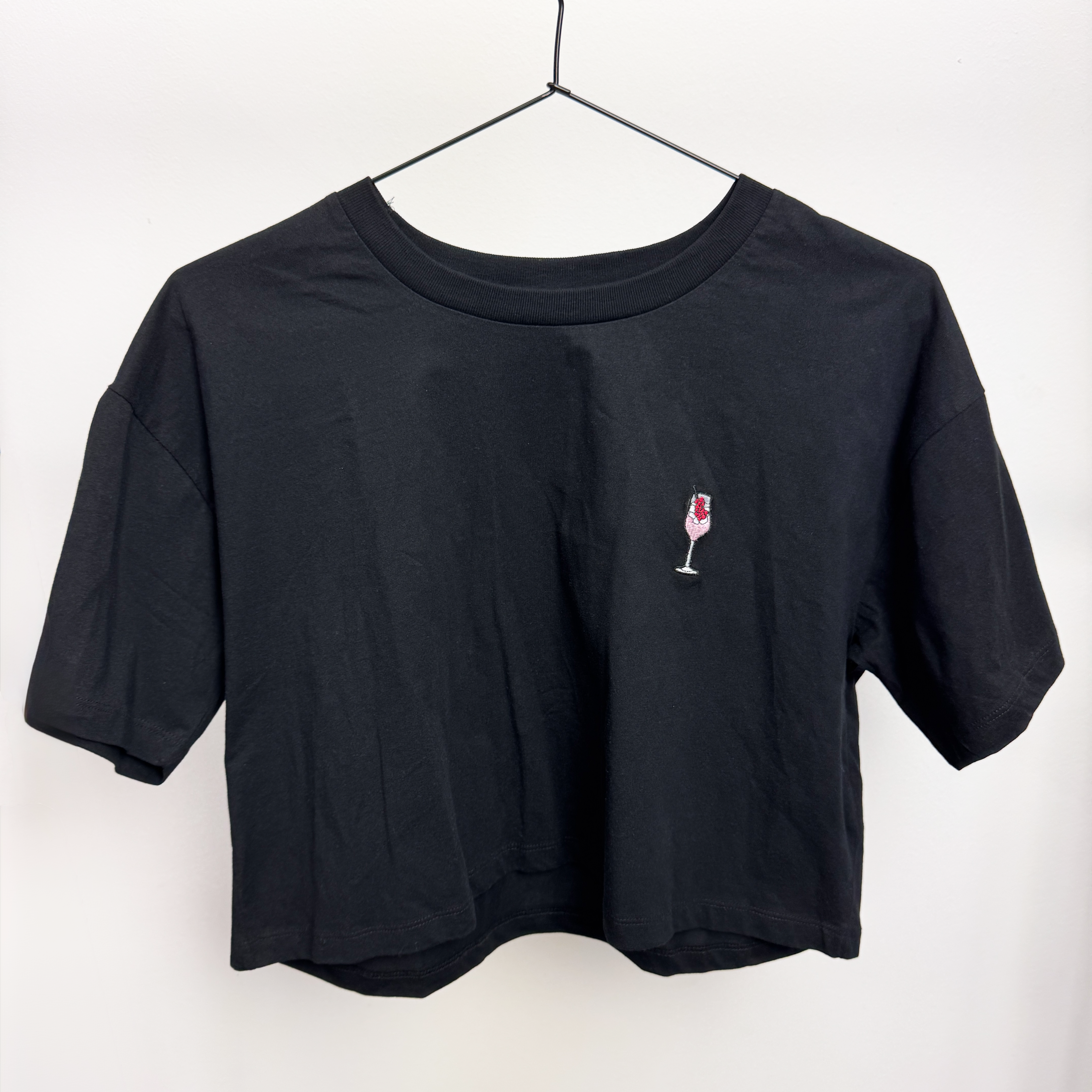 Ein schlichtes schwarzes T-Shirt auf einem Bügel mit einem kleinen gestickten Weinglas mit Rotwein auf der linken Brustseite.
