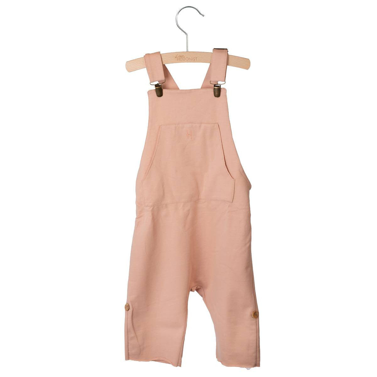 Hellrosa Latzhose für Kleinkinder mit einer Vordertasche, verstellbaren Schulterträgern und seitlichen Knöpfen. Sie hängt an einem Holzbügel vor einem weißen Hintergrund.