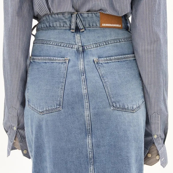 Eine Person trägt ein blaues Nadelstreifenhemd, das in einen hochtaillierten, hellblauen Jeansrock mit sichtbaren Gesäßtaschen und einem ARMEDANGELS-Lederaufnäher am Bund gesteckt ist.