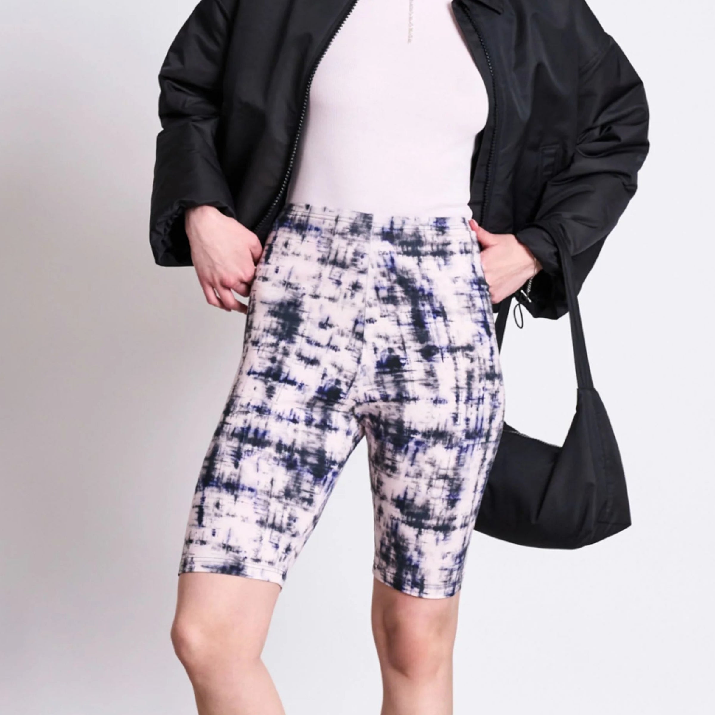 Eine Frau, die eine schwarze Sonnenbrille, eine schwarze Jacke, ein hellrosa Oberteil, gemusterte Radlerhosen, schwarze Plateausandalen und eine kleine schwarze Umhängetasche trägt, steht selbstbewusst vor einem schlichten weißen Hintergrund.