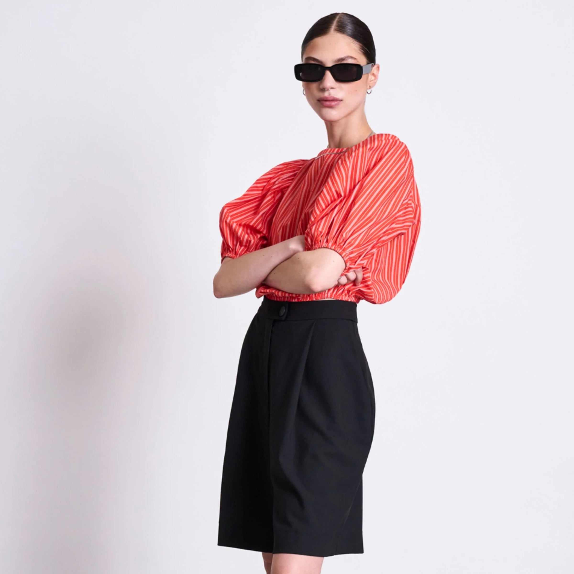 Eine Frau mit Sonnenbrille trägt eine rot gemusterte Bluse mit Puffärmeln, hochgeschnittene schwarze Shorts, durchsichtige schwarze Kniestrümpfe und schwarze Slipper und steht vor einem schlichten weißen Hintergrund.