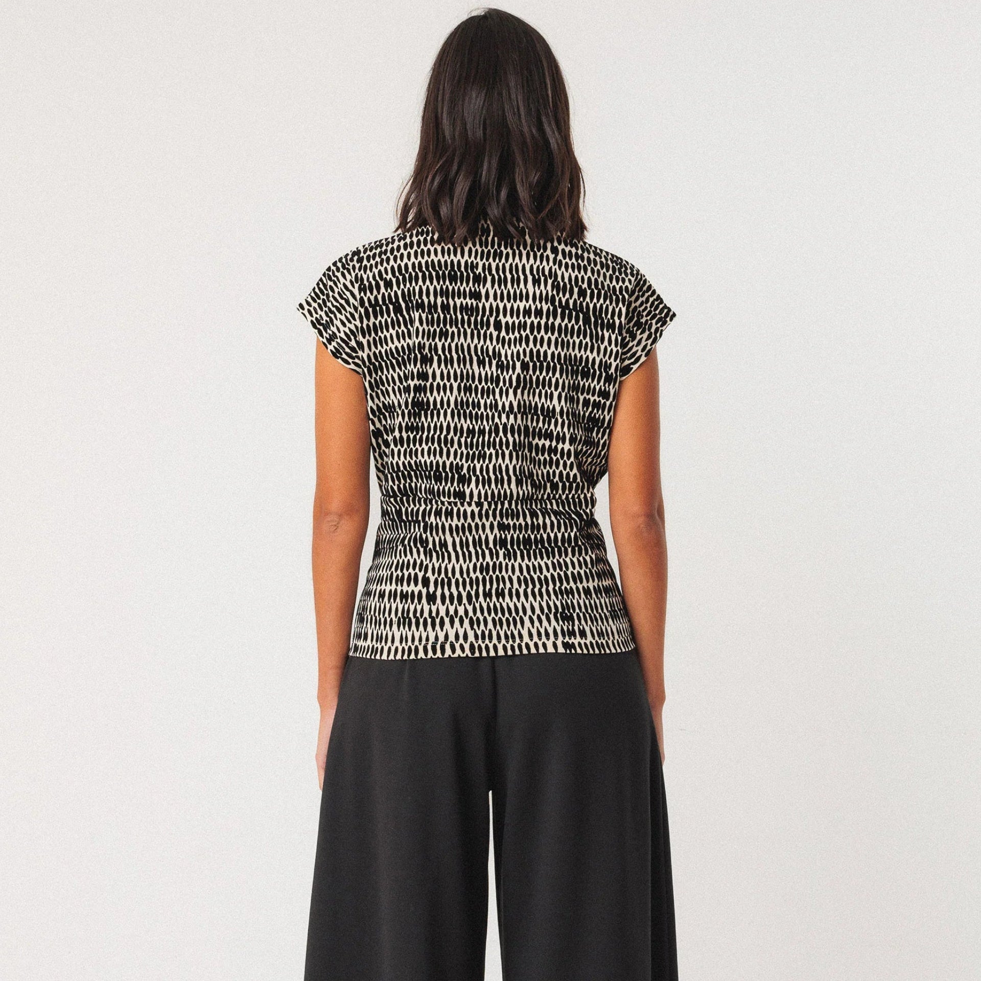 Eine Person steht mit dem Gesicht nach außen, trägt ein kurzärmeliges, gemustertes schwarz-weißes Oberteil und eine schwarze Hose mit weitem Bein und schwarzen Schuhen vor einem schlichten, hellen Hintergrund.