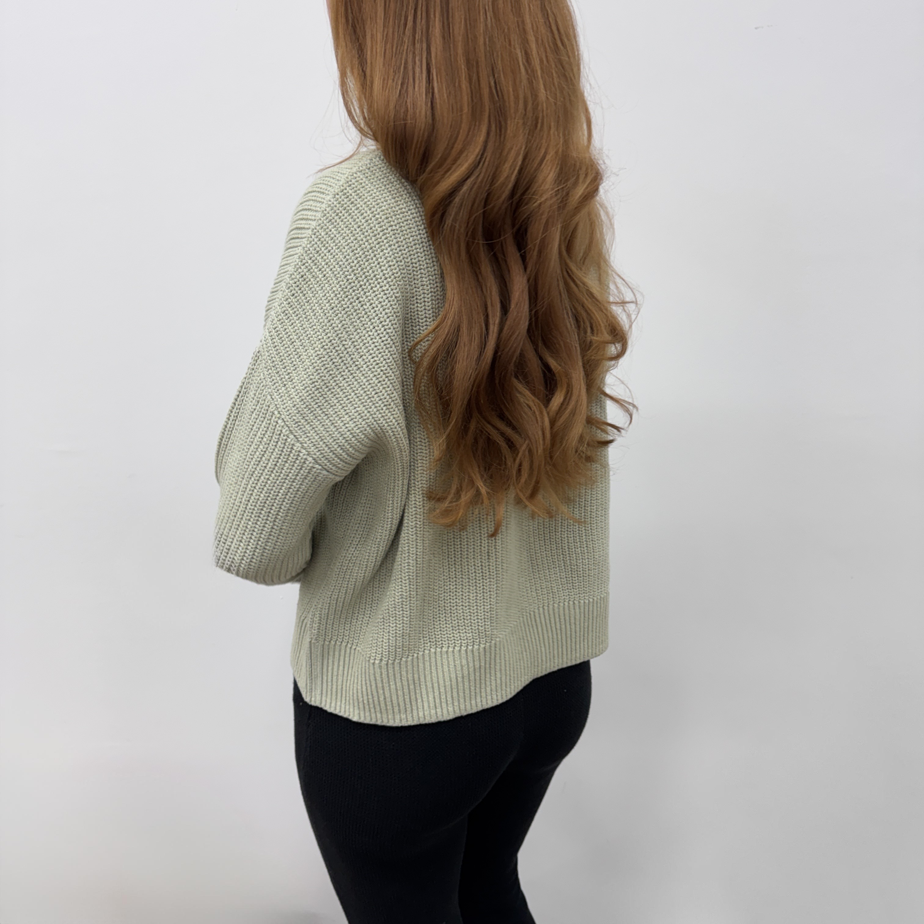 Eine Person mit langem, gewelltem, hellbraunem Haar steht mit dem Gesicht zur Seite. Sie trägt einen hellen Strickpullover und eine dunkle Hose vor einem schlichten weißen Hintergrund.