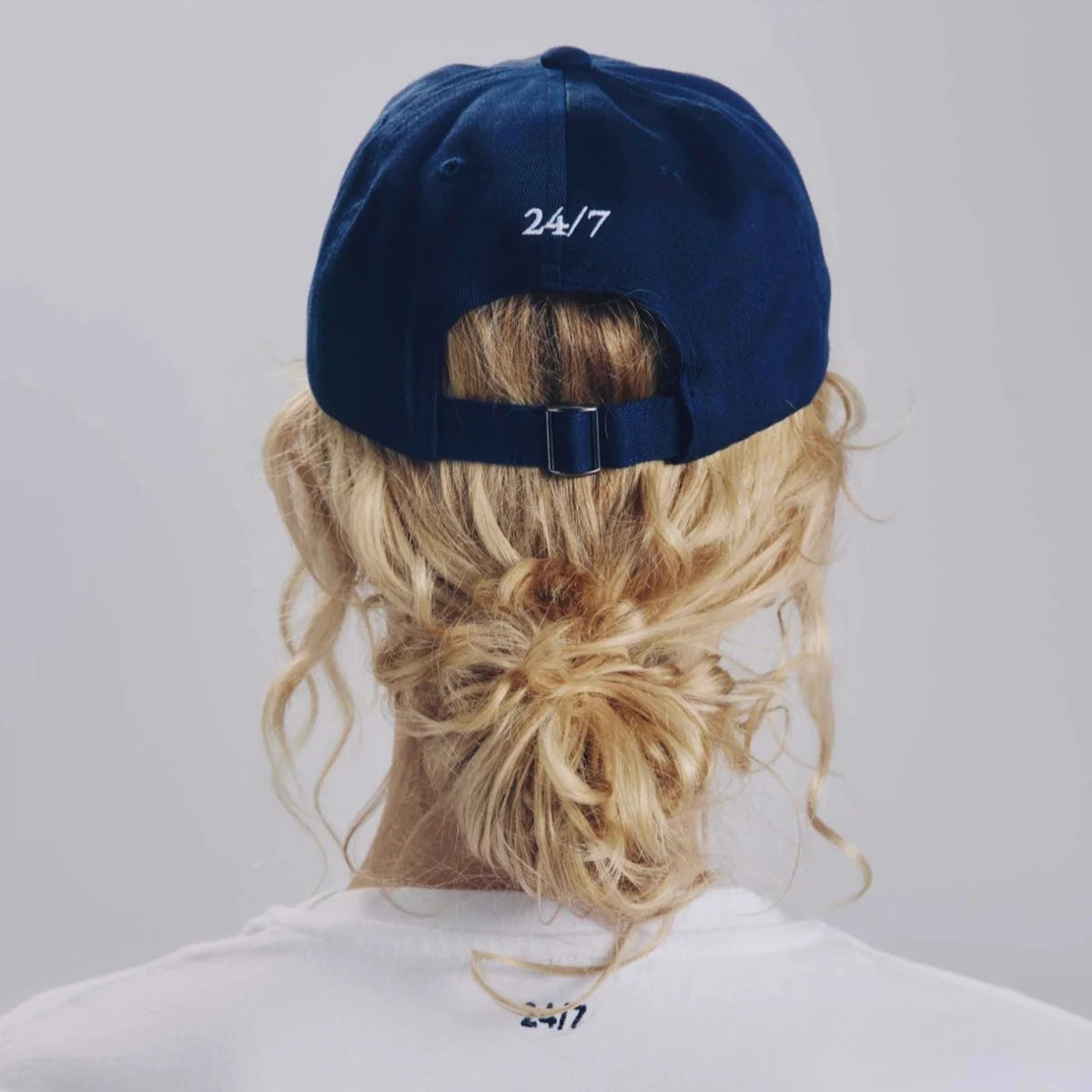 Eine Person mit blondem, gelocktem Haar in einem niedrigen, unordentlichen Dutt trägt eine marineblaue Baseballmütze mit der Aufschrift 24/7 auf der Rückseite. Die Person trägt außerdem ein weißes Hemd mit dem Aufdruck 24/7 am Halsausschnitt. Der Hintergrund ist einfarbig.