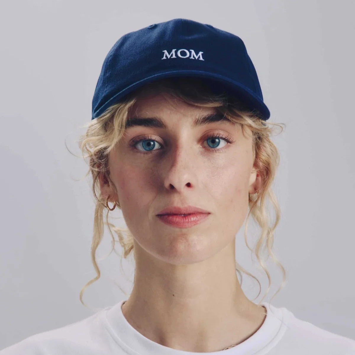 Eine Person mit gewelltem blondem Haar, die eine marineblaue Mütze mit der Aufschrift MOM in weißen Buchstaben und ein schlichtes weißes T-Shirt trägt und vor einem neutralen Hintergrund direkt in die Kamera schaut.