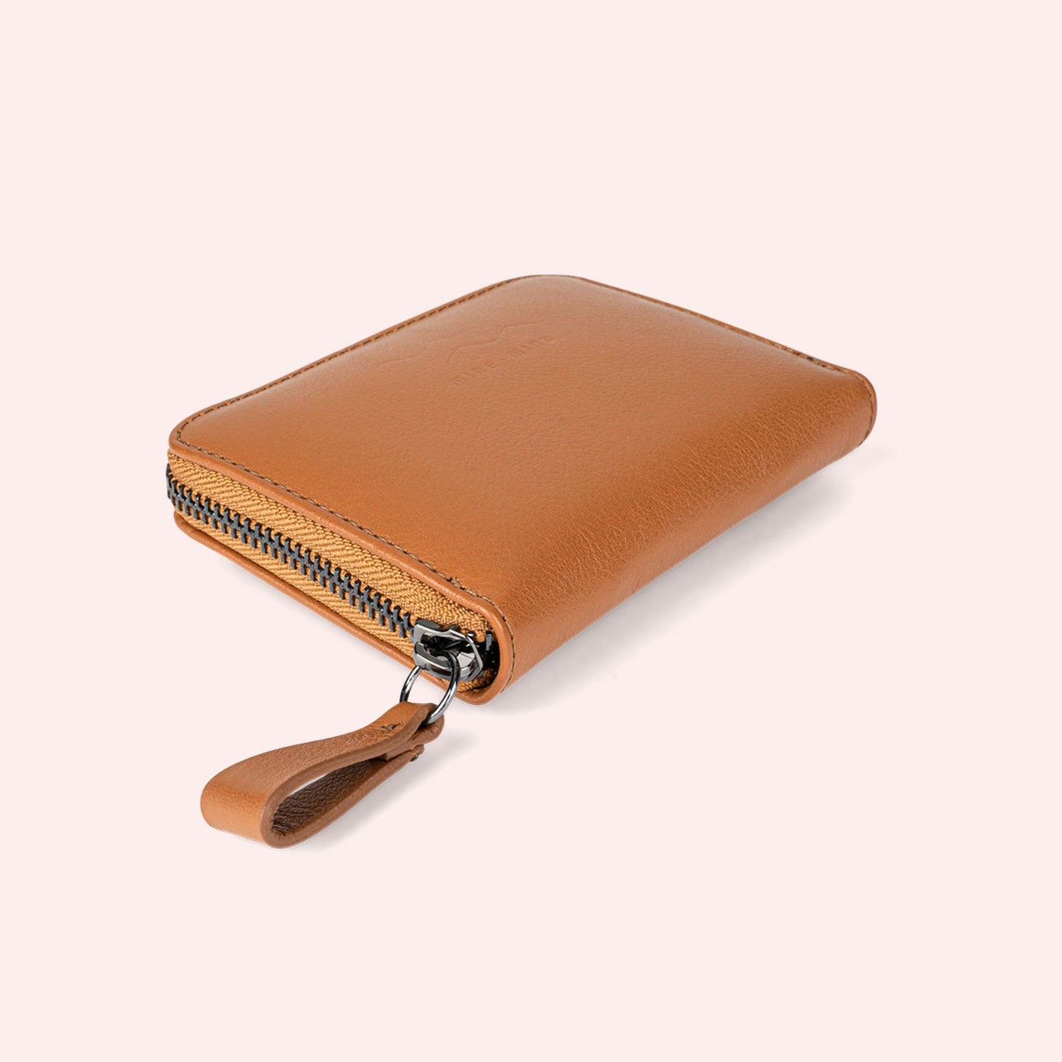 Braunes Lederportemonnaie mit Reißverschluss und passender Lederlasche, platziert auf einem hellrosa Hintergrund. Die Brieftasche ist rechteckig und schräg dargestellt.