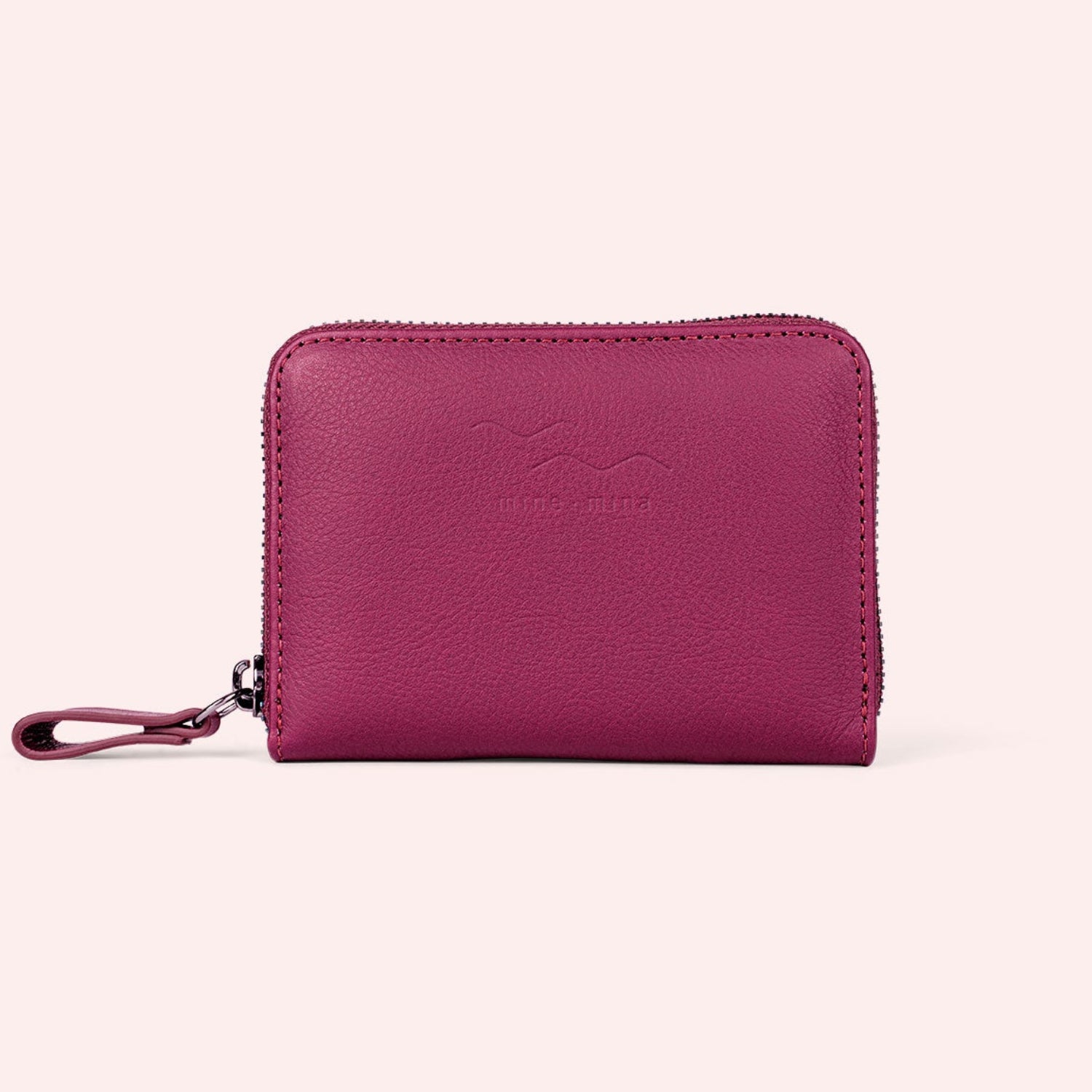 Ein kleines magentafarbenes Lederportemonnaie mit Reißverschluss und einer Handschlaufe, das vor einem hellrosa Hintergrund präsentiert wird.