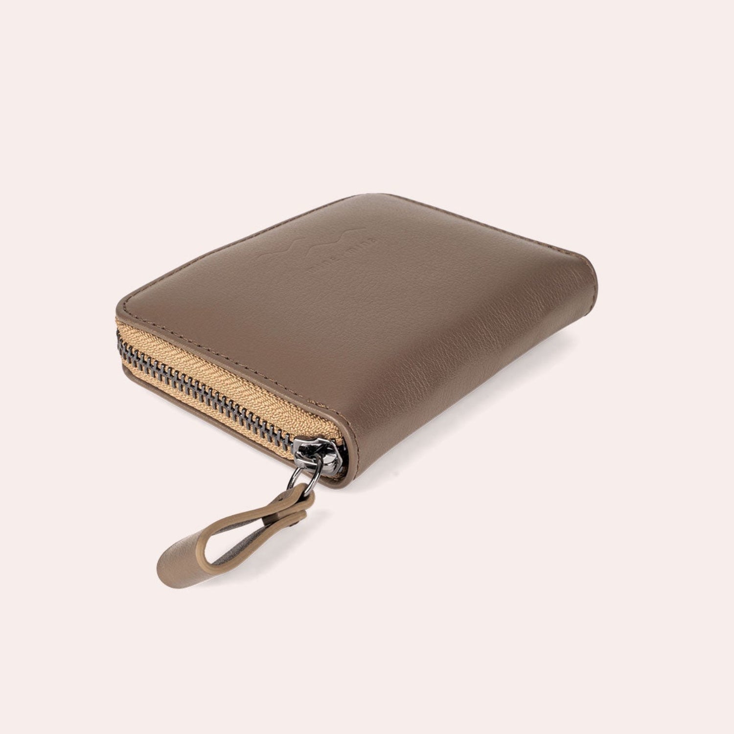 Ein braunes Lederportemonnaie mit Reißverschluss und einer passenden Lederlasche, fotografiert auf einem hellbeigen Hintergrund.
