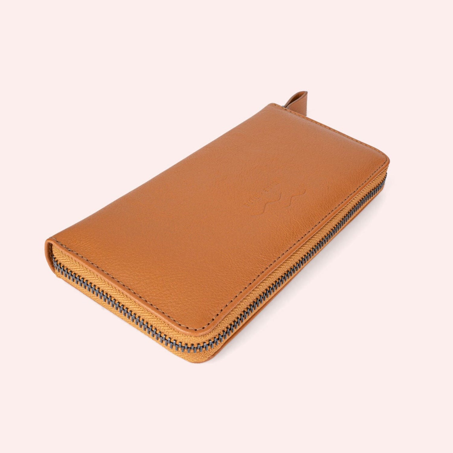 Ein hellbraunes Lederportemonnaie mit Reißverschluss und strukturierter Oberfläche, geschlossen und angewinkelt auf einem hellrosa Hintergrund dargestellt. Der Reißverschluss ist dunkel und hat eine Zuglasche aus Leder.